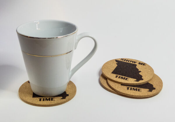 Cork coasters, engraved with the state of Missouri shape and the words "Show Me Time". 3 stacked and one under a glass mug.