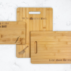 Three Bamboo cutting boards of different sizes. Largest has handle cutout and is engraved with a meat cleaver and the words "On the chopping block" Medium board has a handle cutout and is engraved with the words "A cut above the rest" smallest board is engraved with chilli peppers and the words "Chop it like it's hot"