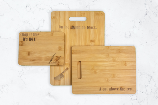 Three Bamboo cutting boards of different sizes. Largest has handle cutout and is engraved with a meat cleaver and the words "On the chopping block" Medium board has a handle cutout and is engraved with the words "A cut above the rest" smallest board is engraved with chilli peppers and the words "Chop it like it's hot"