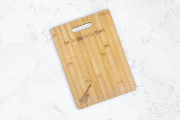 Bamboo cutting board with handle cutout, engraved with a meat cleaver in the bottom left corner, and the words "On the chopping block" at the top, under the handle.