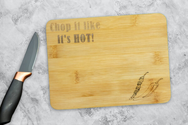 Small Bamboo cutting board, engraved with the words "Chop it like it's hot" in the top-left corner, and 2 chili peppers in the bottom-right, one is chopped. A knife is laying next to it.