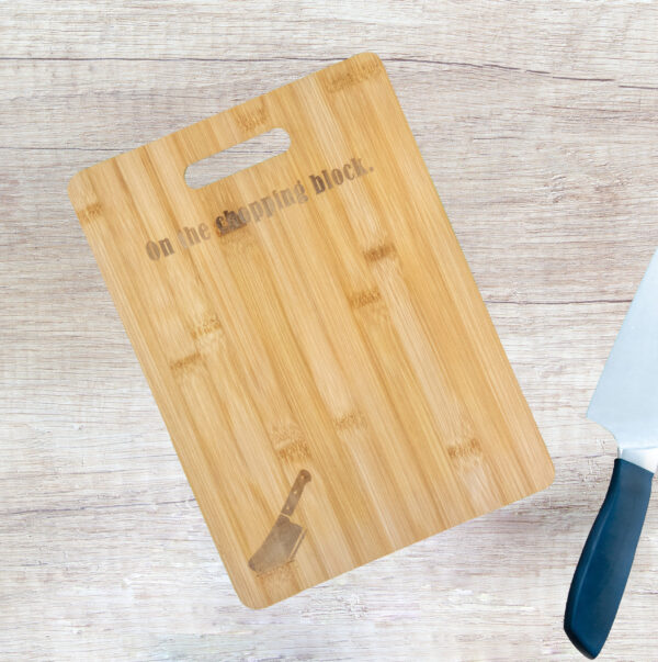 Bamboo cutting board with handle cutout, engraved with a meat cleaver in the bottom left corner, and the words "On the chopping block" at the top, under the handle. A knife is lying next to it.