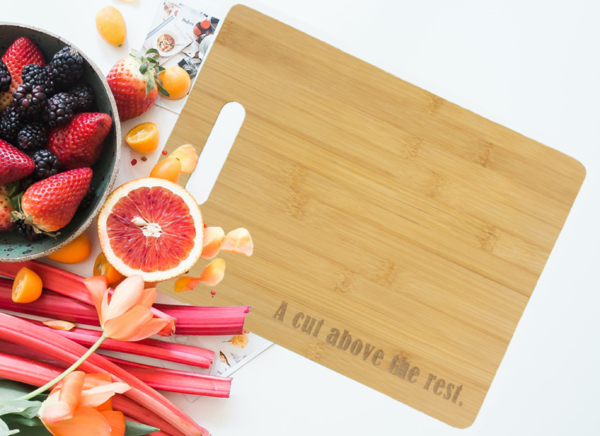 Bamboo cutting board with handle cutout on left side, engraved with the words "A cut above the rest" at the bottom-right. Board is sitting on a counter, surrounded by fruit.