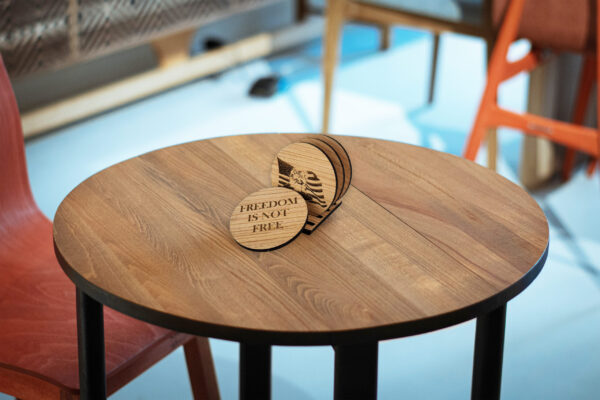 Wooden coasters on a table. Three in the stand, visible one engraved with the head of a bald eagle in front of the American flag. Coaster leaning against the stand says "Freedom is not free"