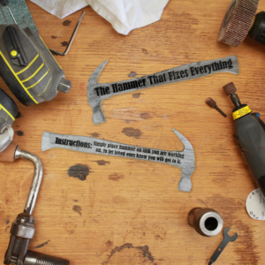 Both sides of a wooden hammer shape that is painted silver and engraved with the words "Instructions: Simply place hammer on task you are working on, to let loved ones know you will get to it." on one side and "The hammer that fixes everything" on the other side. Both are laying on a workbench, surrounded by various tools.