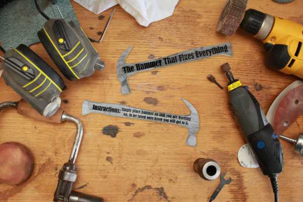 Both sides of a wooden hammer shape that is painted silver and engraved with the words "Instructions: Simply place hammer on task you are working on, to let loved ones know you will get to it." on one side and "The hammer that fixes everything" on the other side. Both are laying on a workbench, surrounded by various tools.