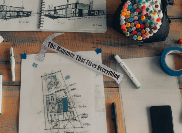 Wooden hammer shape that is painted silver and engraved with the words "The hammer that fixes everything" laying on a desk with project drawings.