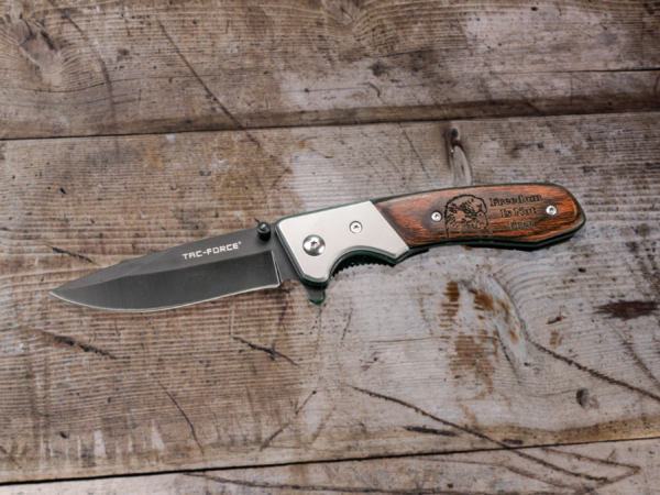 Tac-force pocket knife lying open on a wood table. The wooden handle is engraved with the head of a bald eagle and the words "Freedom is not free"