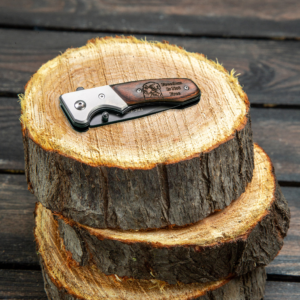 Tac-force pocket knife lying closed on a stack of log slices. The wooden handle is engraved with the head of a bald eagle and the words "Freedom is not free"