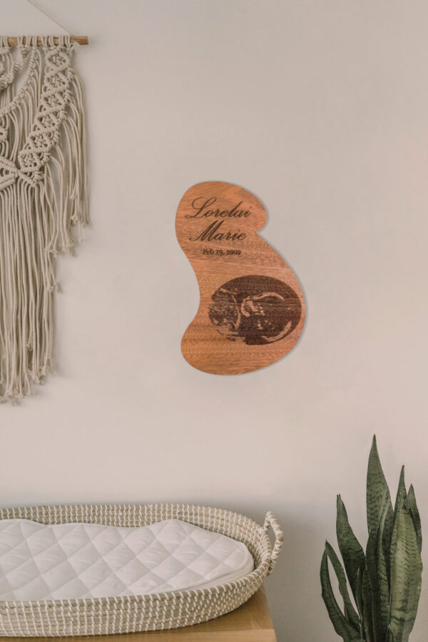 Stylized pregnant bust shaped wooden plaque engraved with an ultrasound image, name, and date, hanging on the wall above a baby changing table.