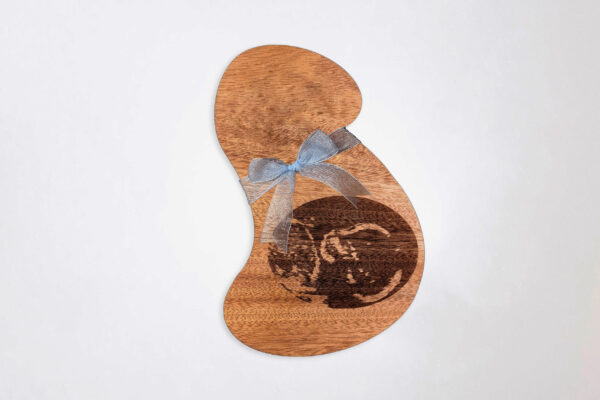 Stylized pregnant bust shaped wooden plaque, engraved with an ultrasound image and tied with a blue ribbon, on a white background.