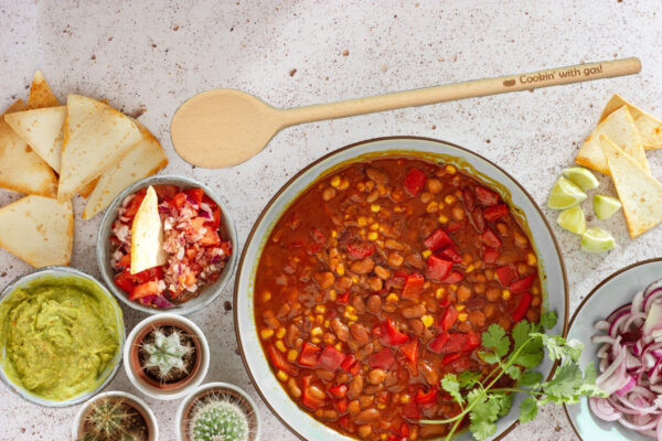 Wood spoon with the words "Cookin' with gas" engraved on the handle laying next to a bowl of chili