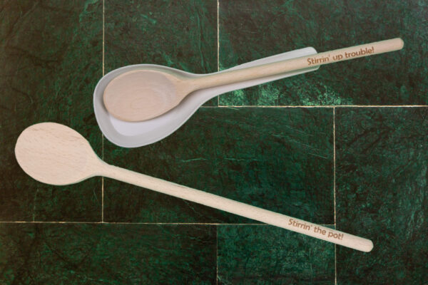 Wood spoon with the words "Stirrin' up trouble" on a spoon rest, next to a wood spoon with the words "Stirrin' the pot"