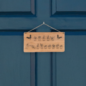 Closeup of a wooden sign laser engraved with ASL handshapes, fingerspelling: Merry Christmas, with two holly sprigs. Sign is hung on a blue door.