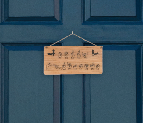 Closeup of a wooden sign laser engraved with ASL handshapes, fingerspelling: Merry Christmas, with two holly sprigs. Sign is hung on a blue door.