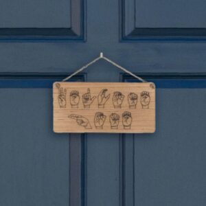 Closeup of a wooden sign laser engraved with ASL handshapes, fingerspelling: Welcome home. Sign is hung on a blue door.