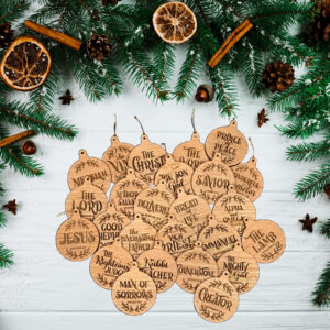 25 Wooden Christmas ornaments, featuring the names of Jesus Christ, laid out on a white table, surrounded by pine boughs.