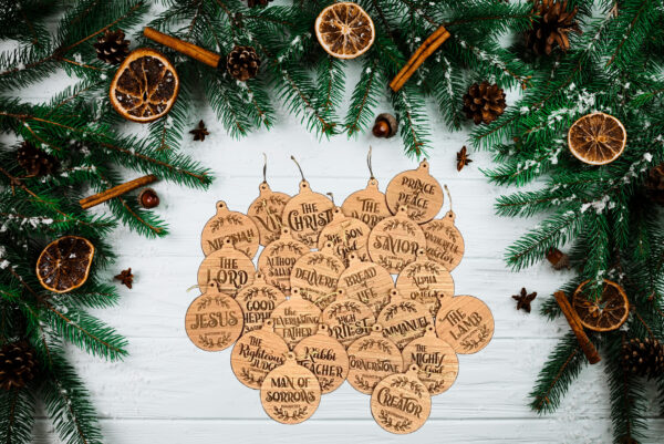 25 Wooden Christmas ornaments, featuring the names of Jesus Christ, laid out on a white table, surrounded by pine boughs.