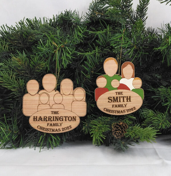 Two wooden family group ornaments hanging on pine boughs. One ornament is painted and features five characters and the words: The Smith Family, Christmas 2022. The second ornament is unpainted and features six characters and the words: The Harrington Family, Christmas 2022