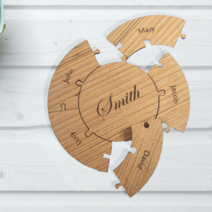 Round wooden puzzle, laser engraved with the name smith on the center piece. 5 edge pieces surround the central piece, each engraved with a first name. Puzzle is partially assembled on a white table, next to a bowl of cherries.