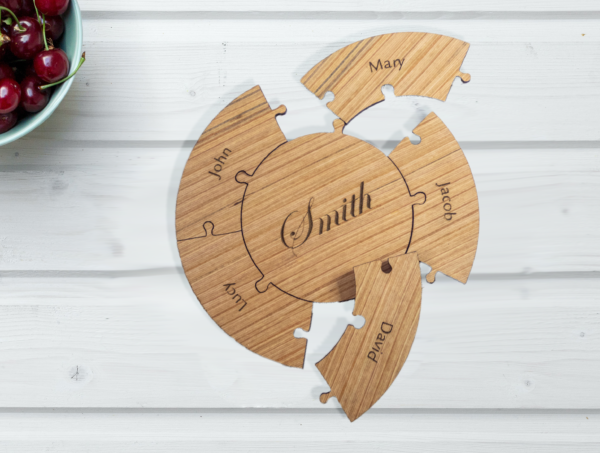 Round wooden puzzle, laser engraved with the name smith on the center piece. 5 edge pieces surround the central piece, each engraved with a first name. Puzzle is partially assembled on a white table, next to a bowl of cherries.