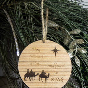 Round wooden ornament, laser engraved with a simple 3 wise men shape under a stylized star and the words "They followed a star... and found a king". Ornament is tied with a jute twine loop and hung on a pine bough.