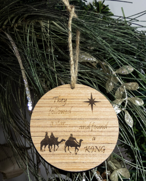 Round wooden ornament, laser engraved with a simple 3 wise men shape under a stylized star and the words "They followed a star... and found a king". Ornament is tied with a jute twine loop and hung on a pine bough.