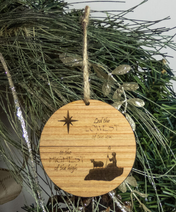 Round wooden ornament, laser engraved with a simple shepherd boy and lamb shape with a stylized star and the words "Led the lowest of the low to the highest of the high". Ornament is tied with a jute twine loop and hung on a pine bough.