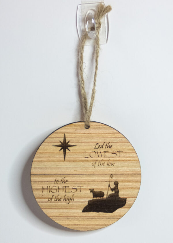 Round wooden ornament, laser engraved with a simple shepherd boy and lamb shape with a stylized star and the words "Led the lowest of the low to the highest of the high". Ornament is tied with a jute twine loop and hung on a white background.