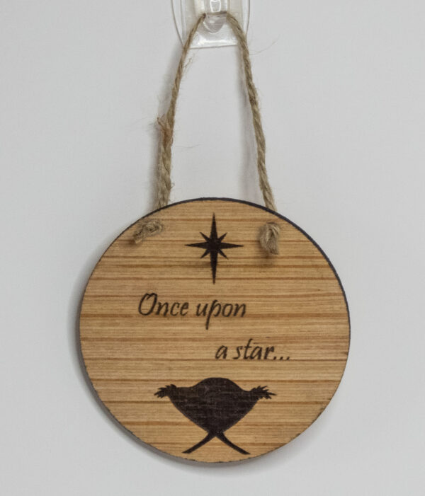 Round wooden ornament, laser engraved with a simple baby in a manger shape under a stylized star and the words "Once upon a star" Ornament is tied with a jute twine loop and hung on a white background.