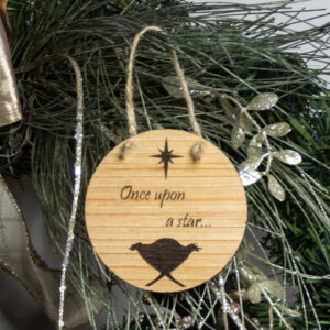 Round wooden ornament, laser engraved with a simple baby in a manger shape under a stylized star and the words "Once upon a star" Ornament is tied with a jute twine loop and hung on a pine bough.