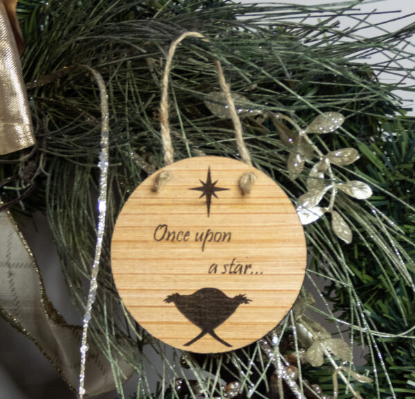 Round wooden ornament, laser engraved with a simple baby in a manger shape under a stylized star and the words "Once upon a star" Ornament is tied with a jute twine loop and hung on a pine bough.