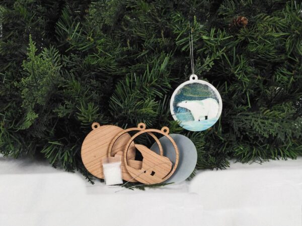 Finished polar bear Christmas snow globe ornament hanging on pine boughs. Ornament kit is sitting next to it.