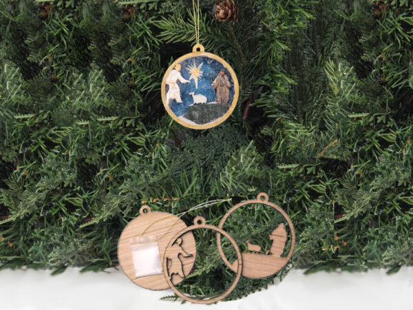 Finished shepherd Christmas snow globe ornament hanging on pine boughs. Ornament kit is sitting next to it.