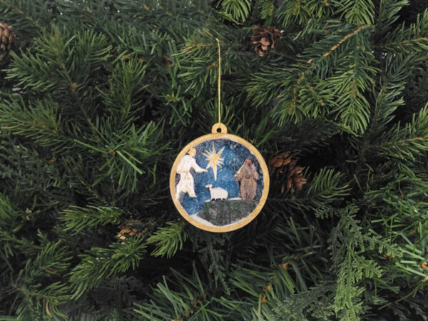 Finished shepherd Christmas snow globe ornament hanging on pine boughs.