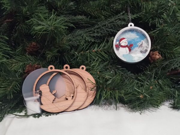 Finished snowman Christmas snow globe ornament hanging on pine boughs. Ornament kit is sitting next to it.