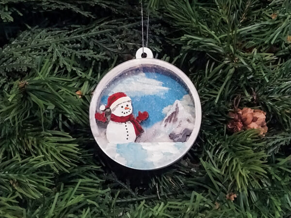 Finished snowman Christmas snow globe ornament hanging on pine boughs.