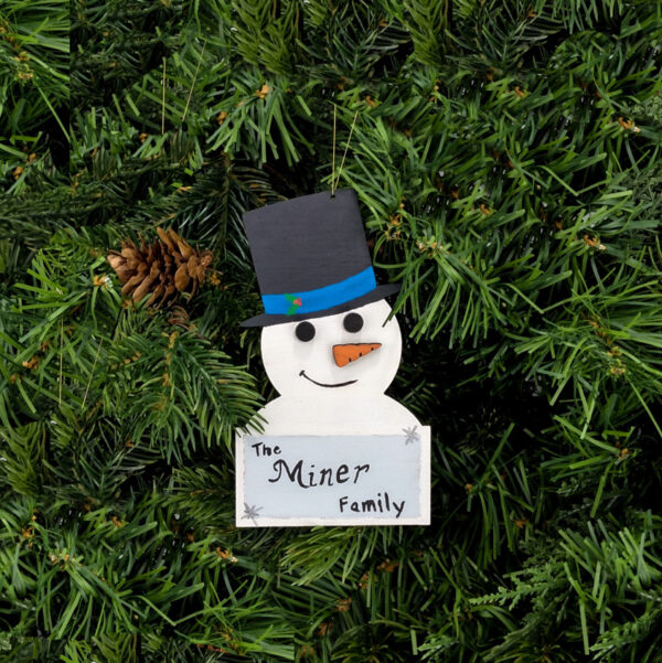 Painted snowman ornament with a top hat and a plaque that says "The Miner Family" hung on pine boughs.