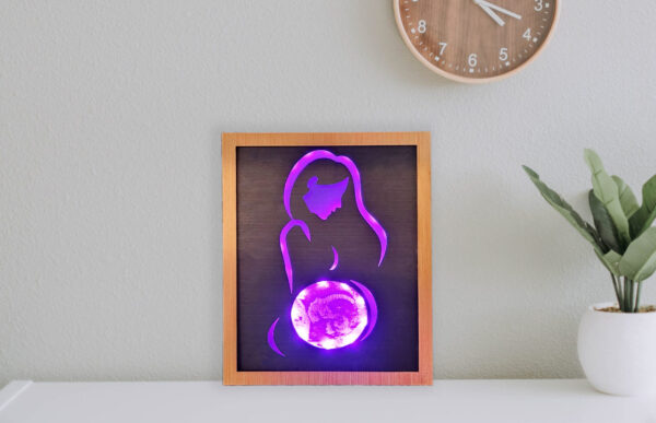 Framed LED lightbox with stylized line silhouette of a woman, the belly displays an ultrasound image engraved on acrylic. Lightbox is illuminated with bright purple light and displayed on a shelf.