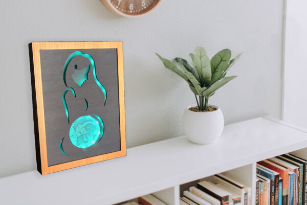 Framed LED lightbox with stylized line silhouette of a woman, the belly displays an ultrasound image engraved on acrylic. Lightbox is illuminated with bright turquoise light and displayed on a shelf.