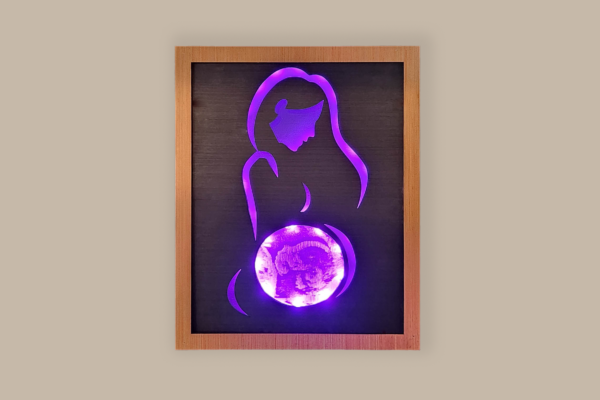 Framed LED lightbox with stylized line silhouette of a woman, the belly displays an ultrasound image engraved on acrylic. Lightbox is illuminated with purple light and displayed on a wall.