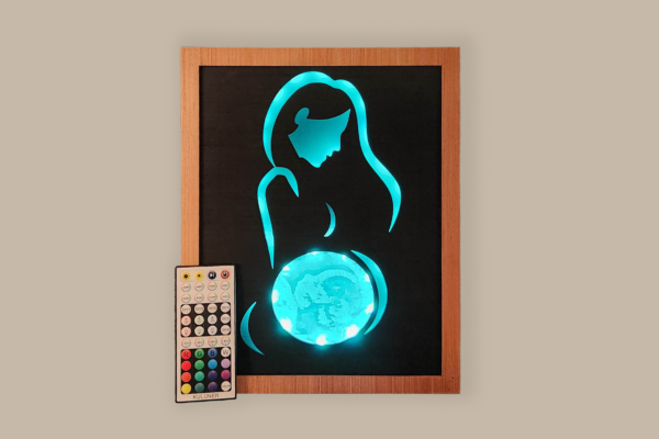 Framed LED lightbox with stylized line silhouette of a woman, the belly displays an ultrasound image engraved on acrylic. Lightbox is illuminated with soft teal light. A remote is displayed at the corner of the piece.