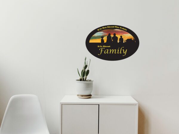 Oval-shaped sign with hand-painted sunset background and silhouette of a family and a cabin. Cutout text says "it is not about the Cabin, it is about family."