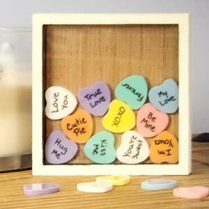 White sweetheart frame with an acrylic face, with colorful acrylic hearts inside with messages written on them, and a few blank hearts laying in front of the frame.