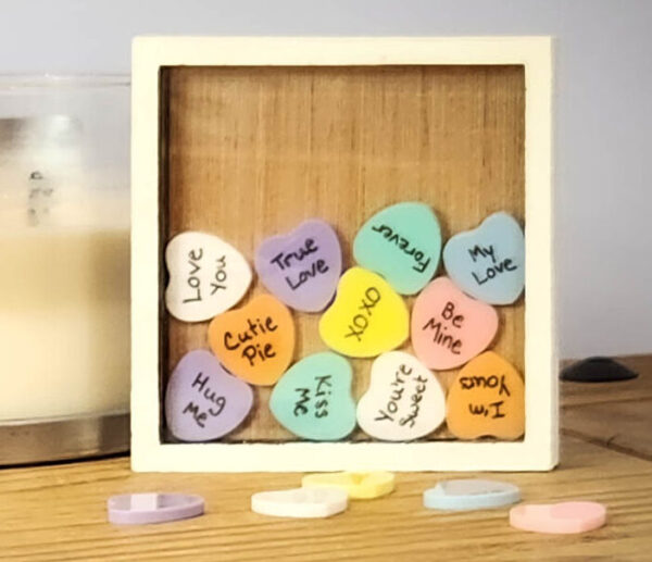 White sweetheart frame with an acrylic face, with colorful acrylic hearts inside with messages written on them, and a few blank hearts laying in front of the frame.