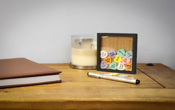 Black sweetheart frame with an acrylic face, with colorful acrylic hearts inside with messages written on them, and a few blank hearts laying in front of the frame with a pen.