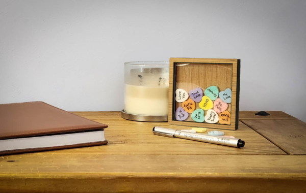 Wood sweetheart frame with an acrylic face, with colorful acrylic hearts inside with messages written on them, and a few blank hearts laying in front of the frame with a pen.